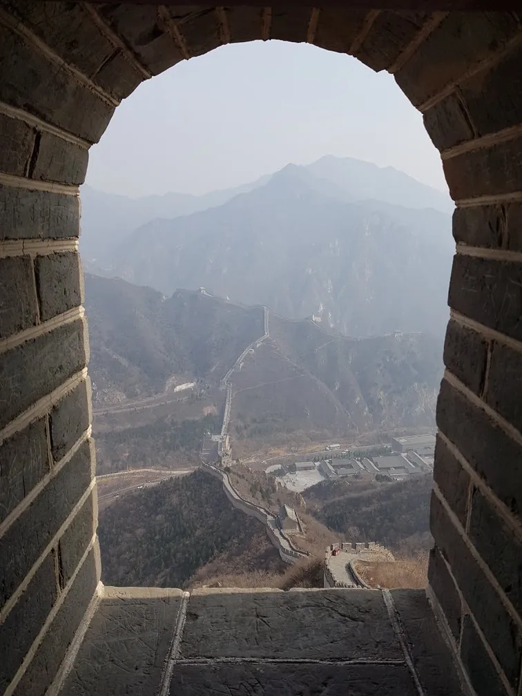 The Great Wall of China framed through the stone arch of a watchtower window, winding down a forested ridge into hazy layered mountains with a village in the valley below.