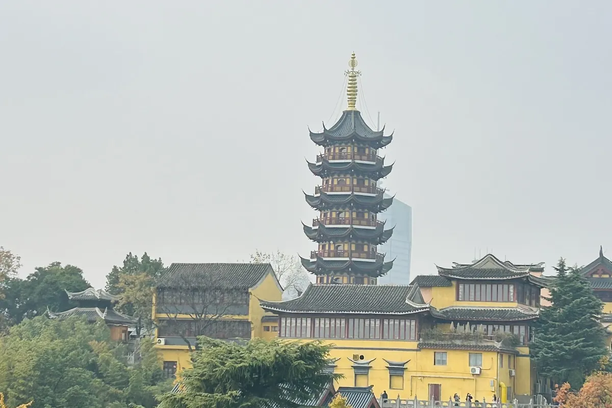 The tiered pagoda of Jiming Temple rising above its yellow halls in Nanjing on a hazy autumn day, with a modern glass skyscraper just visible behind — the old capital's ancient-and-modern blend.