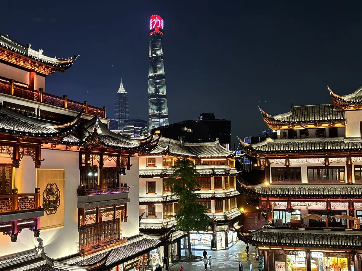 The traditional curved rooftops of Shanghai's Yu Garden bazaar glowing at night, with the Shanghai Tower and Jin Mao Tower rising behind them.