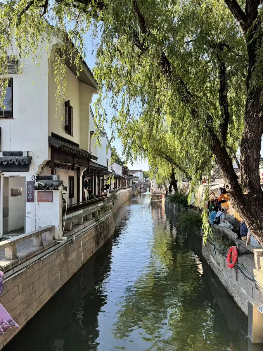 A quiet Suzhou canal lined with whitewashed old houses and weeping willows, a stone arch bridge crossing it in the distance.