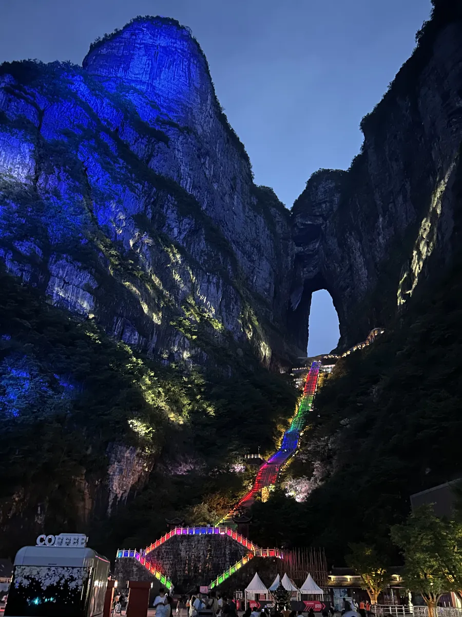 Heaven's Gate — the natural arch of Tianmen Cave — lit in rainbow colours at dusk, the illuminated staircase climbing the cliff to the arch.